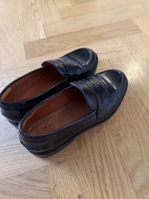 Svarta klassiska loafers i läder - Snygga svarta loafers i glansigt läder med klassisk pennyloafers-design. Skorna har rundad tå, låg klack och bruna innersulor. Perfekta för en stilren och tidlös look. Passar till både jeans och kostymbyxor. Använda endast en gång. Ord. Pris 1500kr