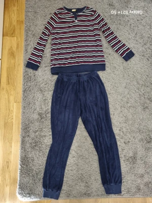 Randig pyjamas från Jolinésse - Mysig tvådelad pyjamas från Jolinésse. Tröjan har marinblå, röda och vita ränder samt knappar vid halsen. Byxorna är enfärgade i mörkblått och har resår i midjan och vid bensluten. Materialet ser ut att vara mjukt och skönt, perfekt för slappa kvällar.