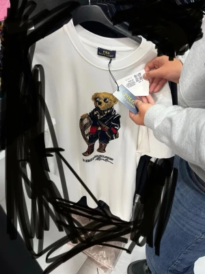 Vit t-shirt med Polo Bear från Ralph Lauren - Vit t-shirt från Polo Ralph Lauren med ikoniskt Polo Bear-motiv på bröstet. Klassisk rund hals och korta ärmar. T-shirten har en avslappnad passform och är tillverkad i mjuk bomull för maximal komfort. Är inte original 