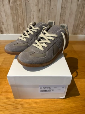Maison Margiela Gats - Snygga grå sneakers med ovandel i mocka och släta läderpaneler. Skorna har vita snören, gummisula och diskreta detaljer för en clean look. Perfekta för dig som gillar stilrena och klassiska sneakers med modern känsla.