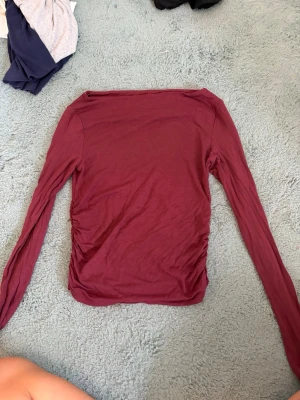Vinröd långärmad topp med rynk - Säljer en vinröd långärmad topp med snygga rynkdetaljer på sidorna. Toppen har en enkel, stilren design och är gjord i ett mjukt bomullsliknande material. Perfekt att matcha med jeans eller kjol för en trendig look.