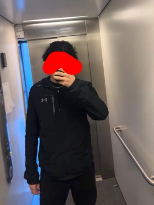 Svart hoodie från Under Armour - Säljer en svart hoodie från Under Armour med dragkedja och huva. Hoodien har en diskret logga på bröstet och är tillverkad i ett sportigt, lätt material som passar perfekt till en aktiv vardag. Snygg och enkel design som funkar till det mesta.