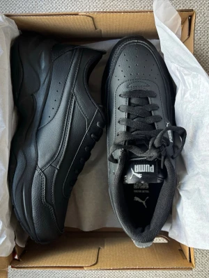 Svarta chunky sneakers från Puma - Stilrena svarta sneakers från Puma med chunky sula och klassisk design. Skorna är tillverkade i syntetläder med perforerade detaljer på ovandelen och har svarta skosnören. Perfekta för dig som gillar en sportig och modern look.