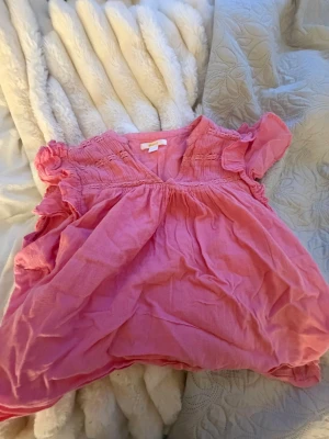 Rosa blus med volang från Gina Tricot - Söt rosa blus från Gina Tricot med volangdetaljer vid ärmarna och smock upptill. Blusen har en luftig passform och V-ringning, perfekt för en avslappnad och trendig look. Tillverkad i ett mjukt, tunt tyg som känns skönt mot huden.