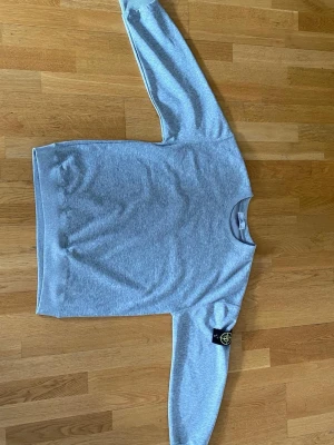Grå sweatshirt från Stone Island - Snygg grå sweatshirt från Stone Island med klassisk rund hals och ribbade muddar. Tröjan har den ikoniska svarta logotypen med gult och grönt broderi på vänster ärm. Perfekt för en avslappnad och trendig stil.