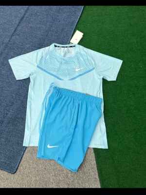 Ljusblått träningsset från Nike - Säljer ett ljusblått träningsset från Nike med t-shirt och shorts. T-shirten har kort ärm, diskret mönster och vit Nike-logga på bröstet. Shortsen är i matchande blå färg med vit logga och elastisk midja. Perfekt för träning eller sport.