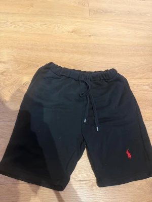 Svarta shorts från Polo Ralph Lauren -  Står M men Passar S. Svarta shorts från Polo Ralph Lauren med dragsko i midjan och den klassiska röda loggan broderad på benet. Tillverkade i mjukt bomullsmaterial för en avslappnad och bekväm stil.