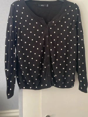 Svart prickig kofta från Ellos - Snygg svart kofta från Ellos med vita prickar och knäppning framtill. Långärmad modell med rund halsringning och ribbade muddar. Perfekt för att addera lite mönster till din outfit.