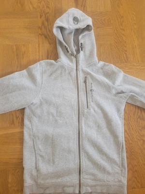 Ljusgrå sail racing zip hoodie - Säljer en ljusgrå hoodie med hel dragkedja framtill och huva med dragsko. Tröjan har en bröstficka med dragkedja och tryckt logga på bröstet. Tillverkad i mjukt sweatshirtmaterial och har ribbade muddar vid ärmslut och nederkant.