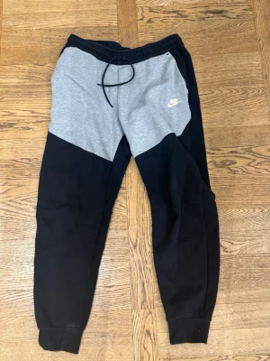 Nike mjukisbyxor svart och grå - Snygga mjukisbyxor från Nike i svart och grått med dragsko i midjan och ribbade muddar vid bensluten. Byxorna har en modern passform och Nike-logga på vänster sida. Perfekta för chill eller träning.