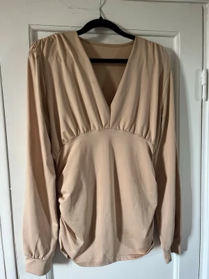 Beige blus med v-ringning - Stilren beige blus med djup v-ringning och rynkade detaljer vid axlar och midja. Blusen har långa ärmar med breda muddar och är tillverkad i ett mjukt, lätt material som ger ett snyggt fall. Perfekt för dig som gillar en enkel men trendig look.