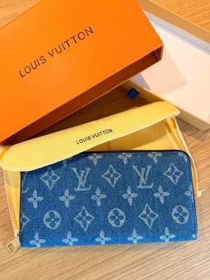 Blå trendig, modern, klassisk plånbok el clutch i denim  - Snygg blå plånbok el clutch i denim med ikoniskt monogrammönster över hela ytan. Plånboken har dragkedja i metall och en rektangulär form. Perfekt för dig som vill ha en trendig accessoar med lyxig känsla.