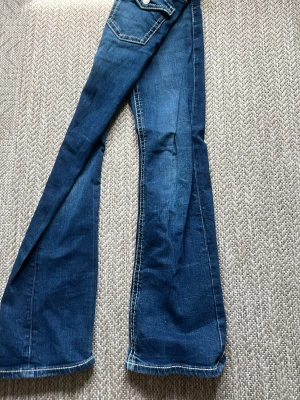 Blå bootcut jeans med kontrastsömmar - Säljer ett par blå bootcut jeans med vita kontrastsömmar och klassiska fickor bak. Jeansen har låg midja och utsvängda ben, vilket ger en retro och trendig vibe. Perfekta för dig som gillar en avslappnad men ändå snygg stil.
