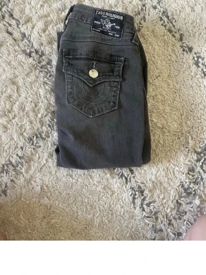 Mörkgrå jeans från True Religion - Säljer snygga mörkgrå jeans från True Religion med klassisk fickdesign och kontrastsömmar. Jeansen har en ikonisk bakficka med lock och silverfärgad knapp. Storlek 25 knappast andvända 