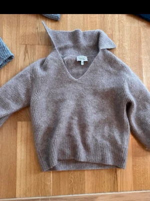 Beige ribbstickad tröja med krage - Mysig beige stickad tröja med bred krage och v-ringning. Tröjan har ribbstickat mönster och långa ärmar. Perfekt för lager på lager och en avslappnad men snygg look.