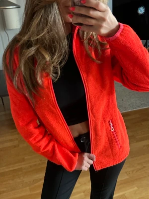 Orange fleecejacka med dragkedja - Säljer en riktigt snygg orange fleecejacka med hel dragkedja framtill och fickor med dragkedja. Jackan har en enkel, sportig look och passar perfekt till vardags eller när du vill hålla dig varm. Mjuk och skön med normal passform och lång ärm.