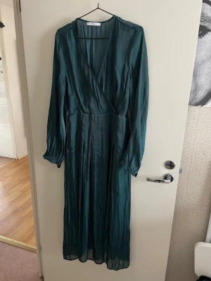 Joelle Mörkgrön glansiga midi klänning med lång ärm, stl 40, svensk design  - Joelle Snygg långklänning i mörkgrönt med omlottmodell och långa ärmar. Klänningen har ett lätt glansigt material och v-ringad hals. Perfekt för dig som vill ha en elegant och stilren look. 
