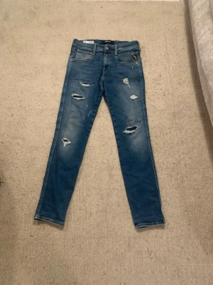 Blå Replay Jeans - Säljer ett par blå skinny jeans med slitningar och hål på framsidan. Jeansen har klassisk femficksdesign, normal midja och dragkedja med knapp. Perfekta för en avslappnad och trendig look.