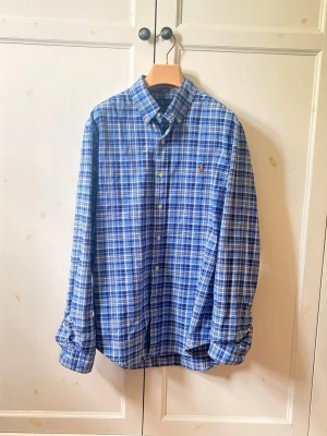 Ralph Lauren skjorta - Klassisk blå och vit rutig skjorta med knappar framtill och button-down krage. Skjortan har lång ärm och en liten broderad detalj på bröstet. Perfekt för en avslappnad men ändå stilren look. 
