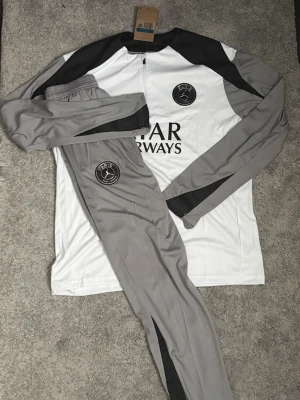 PSGTracksuit  - Säljer en PSG träningsoverall med långärmad tröja i vitt, grått och svart samt matchande grå byxor med svarta detaljer. Setet har PSG-logga och Qatar Airways-tryck. Perfekt för fotbollsträning eller sportiga dagar.