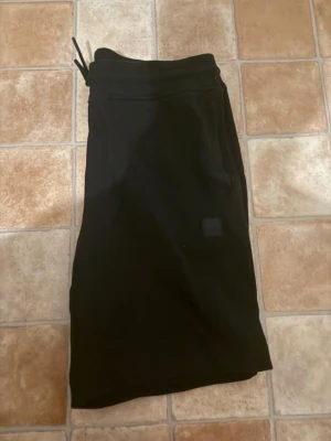 Svarta shorts med snörning - Säljer ett par svarta shorts med snörning i midjan och enkel design. Shortsen har en liten patch framtill och är tillverkade i ett mjukt material som känns skönt mot huden. Perfekta för avslappnade dagar eller träning. Xl men sitter som L