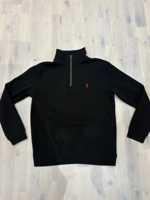 Svart half zip tröja från Polo Ralph Lauren - Svart långärmad tröja från Polo Ralph Lauren med half zip och klassisk röd logga broderad på bröstet. Tröjan har hög krage och är tillverkad i mjukt bomullsmaterial. Perfekt för en stilren och avslappnad look.