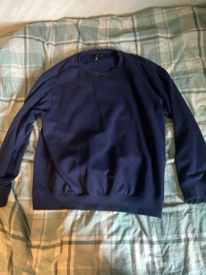 Mörkblå sweatshirt med rund hals - En basic mörkblå sweatshirt med rund halsringning och långa ärmar. Tröjan har ribbade muddar vid ärmslut och nederkant. Perfekt för en avslappnad och stilren look. Materialet ser ut att vara mjukt och bekvämt, troligen bomull eller bomullsblandning.