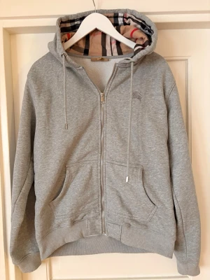 Grå zip hoodie från Burberry junior XL - Snygg grå hoodie från Burberry i storlek junior XL (herrS-M). Klassisk dragkedja framtill, två stora fickor och huva med det ikoniska Burberry-mönstret på insidan. Tillverkad i mjukt material med ribbade muddar vid ärmar och nederkant. Perfekt för en chill och stilren look.