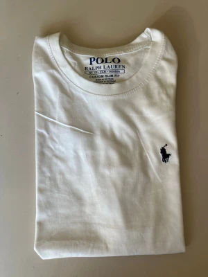 Vit t-shirt från Polo Ralph Lauren - Klassisk vit t-shirt från Polo Ralph Lauren med broderad marinblå logga på bröstet. Modellen är custom slim fit och har rund hals. Tillverkad i mjuk bomull som känns skön mot huden. Perfekt basplagg för en clean och stilren look.