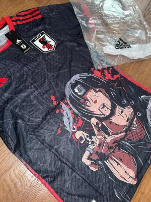 Japan Adidas anime(Naruto) t-shirt svart/röd - Cool svart t-shirt från Adidas med Japan-emblem och röd krage. Perfekt för dig som gillar både fotboll och Itachi. Materialet är lätt och andas.