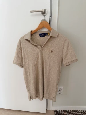 Beige pikétröja från Polo Ralph Lauren - Säljer en beige pikétröja från Polo Ralph Lauren i modellen Custom Slim Fit. Tröjan har korta ärmar, klassisk krage och knappslå framtill. Liten broderad logga på bröstet. Perfekt till en avslappnad men stilren look.