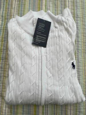 Vit kabelstickad tröja med dragkedja - Snygg vit kabelstickad tröja från Polo Ralph Lauren med dragkedja framtill och broderad marinblå logga på bröstet. Tröjan har ribbade muddar och hög krage, perfekt för en clean och klassisk look.