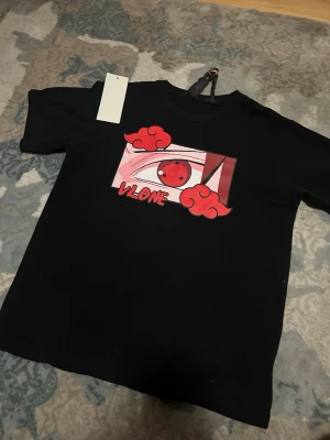 Svart VLONE t-shirt med anime-tryck - Svart t-shirt från VLONE med ett stort anime-inspirerat tryck på bröstet som visar ett rött öga och röda moln. T-shirten har rund hals och korta ärmar. Perfekt för dig som gillar streetwear och anime.