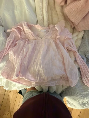 Ljusrosa blus med volanger Gina Tricot - Supersöt ljusrosa blus från Gina Tricot med långa ärmar och volangdetaljer vid axlar och halsringning. Blusen har en luftig, lätt struktur och v-ringning framtill. Perfekt för dig som gillar romantisk och feminin stil.