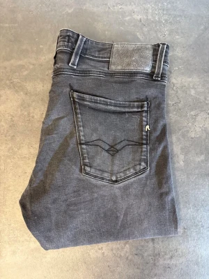 Grå Replay Anbass Jeans, stl 33/34 - Snygga grå jeans i slim fit-modell med klassisk femficksdesign och diskreta slitningar. Jeansen har svarta sömmar och en stilren bakficka med broderad detalj. Perfekta för dig som gillar en enkel men trendig look. NYPRIS:2000kr