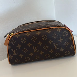 Brun stor, trendig necessär i brunt monogram mönster Helt ny - Säljer en brun necessär med klassiskt monogrammönster i guld. Den är stor och rymlig och trendig modell som håller i år efter år. Bra kvalité. Väskan har två dragkedjor upptill, guldfärgade detaljer och en liten handrem med röda och beige inslag. 
