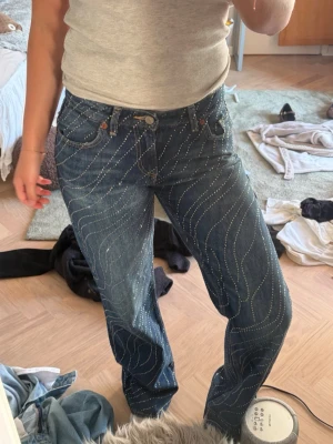 Blå jeans med rhinestones - Säljer ett par blå jeans med raka ben med rhinestones över hela jeansen. Jeansen har klassisk femficksmodell och normal midja. Materialet är denim och de har en riktigt cool och trendig vibe.
