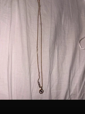 Guldfärgat halsband med pärldetalj Guess - Snyggt guldfärgat halsband från Guess med en rund hängande pärldetalj och tunn kedja. Kommer med en vit påse med Guess-logga. Perfekt accessoar för att lyfta din outfit.