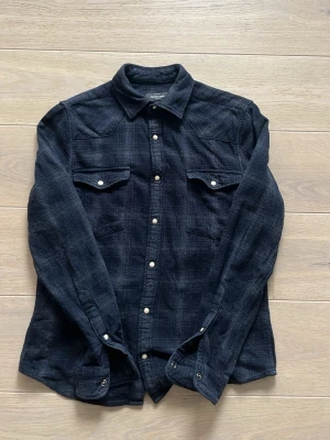Mörkblå rutig skjorta med knappar - Snygg mörkblå overshirt/skjorta med diskret rutigt mönster och vita knappar. Skjortan har två bröstfickor med lock och lång ärm. Perfekt till jeans eller chinos för en avslappnad look.