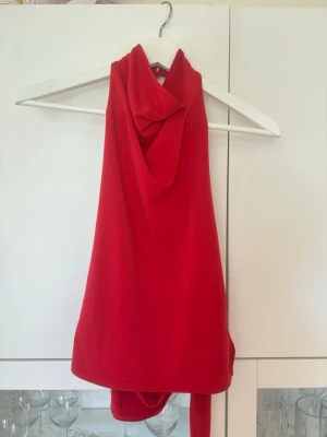 Röd halterneck topp med knyt - Snygg röd halterneck topp med draperad framsida och knyt i nacken. Toppen har en dekorativ ringdetalj på sidan och är ärmlös, vilket ger en trendig och ungdomlig look. Perfekt för dig som vill sticka ut med en färgstark och modern topp.