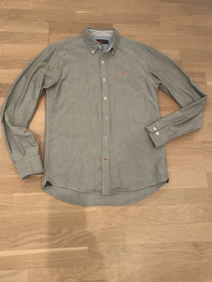 Grå skjorta från Morris - Klassisk grå skjorta från Morris med button down-krage och vita knappar. Skjortan har lång ärm och en liten orange logga broderad på bröstet. Tillverkad i mjuk bomull och har diskreta sömmar för en clean look.