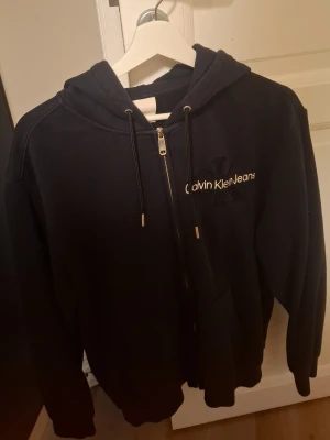Svart zip-hoodie från Calvin Klein Jeans - Snygg svart hoodie med dragkedja från Calvin Klein Jeans. Stor broderad CK-logga och text på bröstet. Huva med dragsko och fickor framtill. Perfekt för en chill och stilren look. STORLEK M Använd ett par gånger men passade inte mig personligen så har mest varit på hyllan. Nypris var ganska saftigt minns jag
