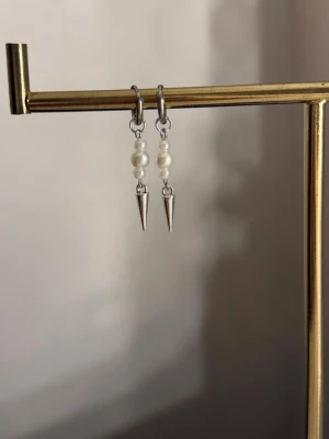 Silvriga örhängen med pärlor och spikes - Säljer ett par trendiga örhängen i silverfärg med två vita pärlor och en spetsig spike-detalj längst ner. Örhängena har en cool kombination av klassiska pärlor och edgy metallspetsar, perfekta för dig som gillar att mixa stilar.