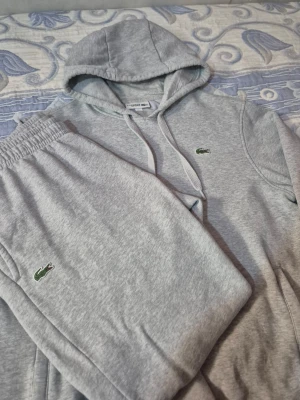 Lacoste Sport tracksuit  - Snygg grå hoodie från Lacoste Sport med klassisk huva och dragsnören. Ikoniska gröna krokodilloggan broderad på bröstet. Tillverkad i mjukt sweatshirtmaterial som känns skönt mot huden. Perfekt för en avslappnad och sportig stil.