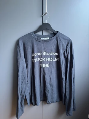 Grå långärmad topp Acne Studios - Säljer en grå långärmad topp från Acne Studios med tryck 'Acne Studios STOCKHOLM 1996' framtill. Toppen har en avslappnad passform och är tillverkad i mjuk bomull, perfekt för en chill och stilren look.