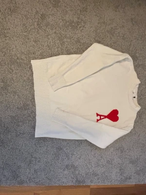 Vit stickad tröja från Ami Paris - Vit stickad tröja från Ami Paris med röd hjärtdetalj och bokstaven A på bröstet. Tröjan har rund halsringning och ribbade muddar. Perfekt för dig som gillar stilrena plagg med en twist. REKOMMENDERA TILL NÅN SOM HAR XS..