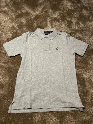 Grå pikétröja från Polo Ralph Lauren - Snygg grå pikétröja från Polo Ralph Lauren i klassisk passform. Tröjan har korta ärmar, krage med knappar och den ikoniska broderade loggan på bröstet. Perfekt för en stilren och avslappnad look.