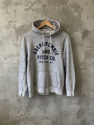 Grå hoodie från Abercrombie & Fitch - Snygg ljusgrå hoodie från Abercrombie & Fitch med mörkblå text framtill. Klassisk känguruficka och justerbar huva med vita snören. Perfekt för en avslappnad och trendig look. Mjuk och skön bomullsblandning.