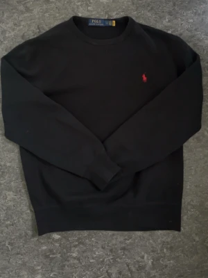 Svart sweatshirt från Polo Ralph Lauren - Klassisk svart sweatshirt från Polo Ralph Lauren med rund halsringning och röd broderad logga på bröstet. Tröjan har långa ärmar och ribbade muddar vid ärmslut och nederkant. Perfekt för en stilren och avslappnad look.