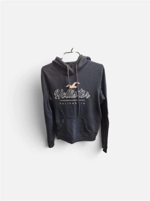 Mörkblå hoodie från Hollister - Snygg mörkblå hoodie från Hollister med stor logga och texten 'California' på bröstet. Hoodien har huva med dragsko och en klassisk magficka. Perfekt för en avslappnad och trendig look.